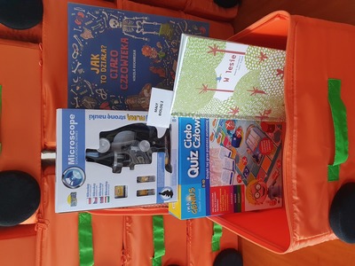Na zdjęciu znajduje się pomarańczowa otwarta walizeczka. W jej środku znajduje się gra edukacyjna, książka, gadżet edukacyjny..
