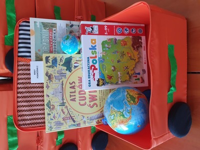 Na zdjęciu znajduje się pomarańczowa otwarta walizeczka. W jej środku znajduje się gra edukacyjna, książka, gadżet edukacyjny..