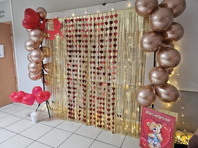 Na zdjęciu aranżacja z różowymi balonami, czerwonymi sercami i złotymi paskami zawieszonymi na stelażu 