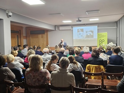 Duża grupa ludzi siedzących na krzesłach na sali i patrzących na przemawiającego mężczyznę