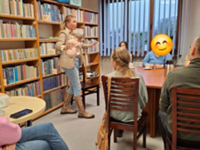 Spotkanie w pomieszczeniu bibliotecznym, regały z ksiązkami, prowadząca stoi trzyma lalkę niemowlęcia, osoby dorosłe dziedzą przy stole