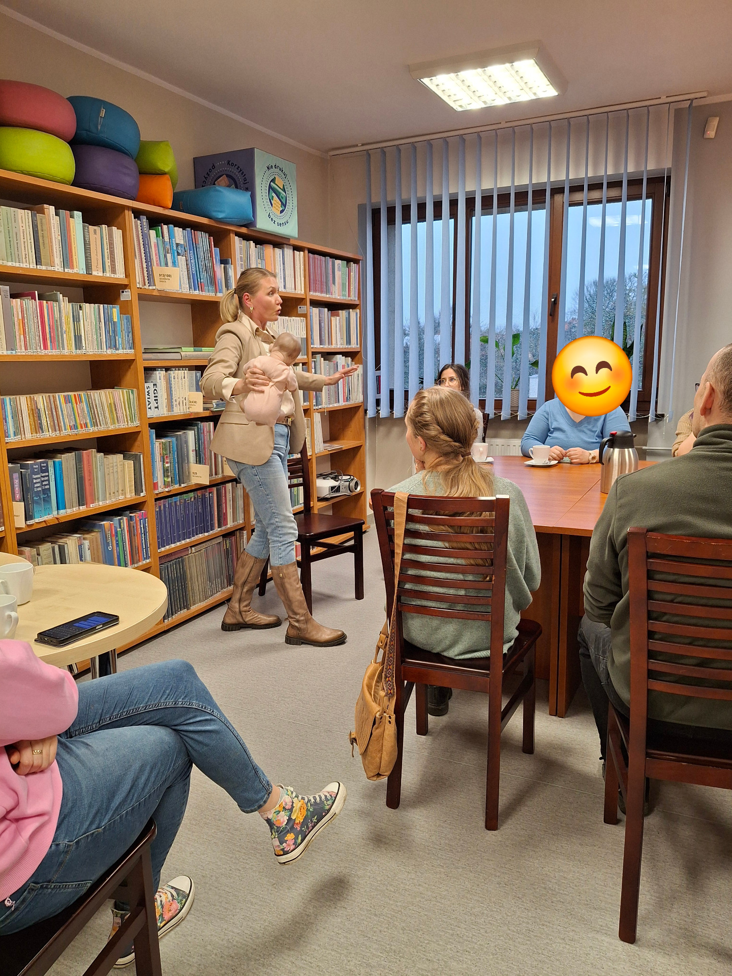 Spotkanie w pomieszczeniu bibliotecznym, regały z ksiązkami, prowadząca stoi trzyma lalkę niemowlęcia, osoby dorosłe dziedzą przy stole