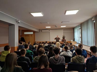 publiczność szkolna siedząca tyłem, na przodzie strój wikinga na wieszaku oraz prowadzący mężczyzna w dawnym stroju