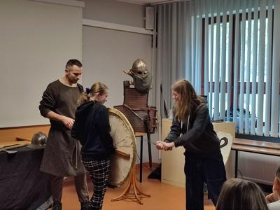 dwie dziewczyny jedna trzyma tarcze, druga miecz, obok stoi mężczyzna w dawnym stroju, w tle zbroja wikinga na wieszaku