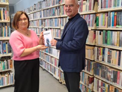 kobieta w różowym swetrze, czarnych spodniach odbiera książkę od mężczyzny w granatowej marynarce i czarnych spodniach, w tle regały biblioteczne