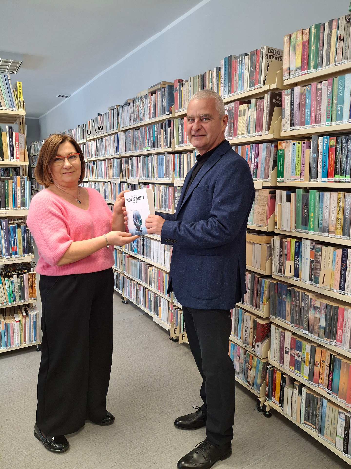 kobieta w różowym swetrze, czarnych spodniach odbiera książkę od mężczyzny w granatowej marynarce i czarnych spodniach, w tle regały biblioteczne