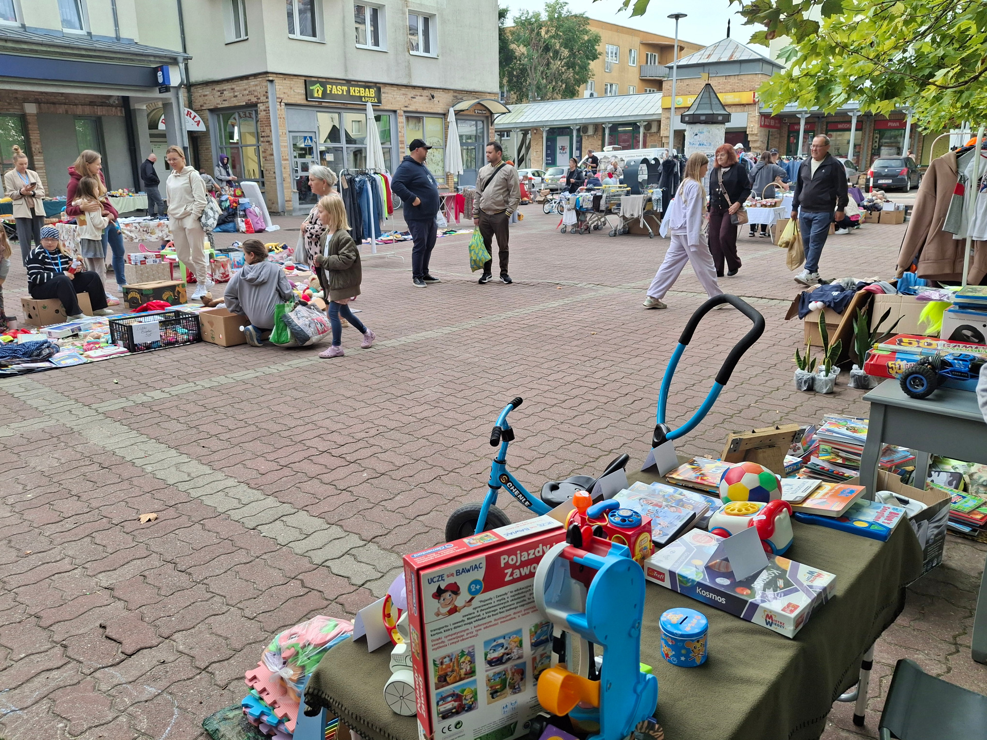 rynek w mieście, stoiska pchli targ, ludzie pomiedzy nimi, na stoiskach zabawki, ubrania, rowerki