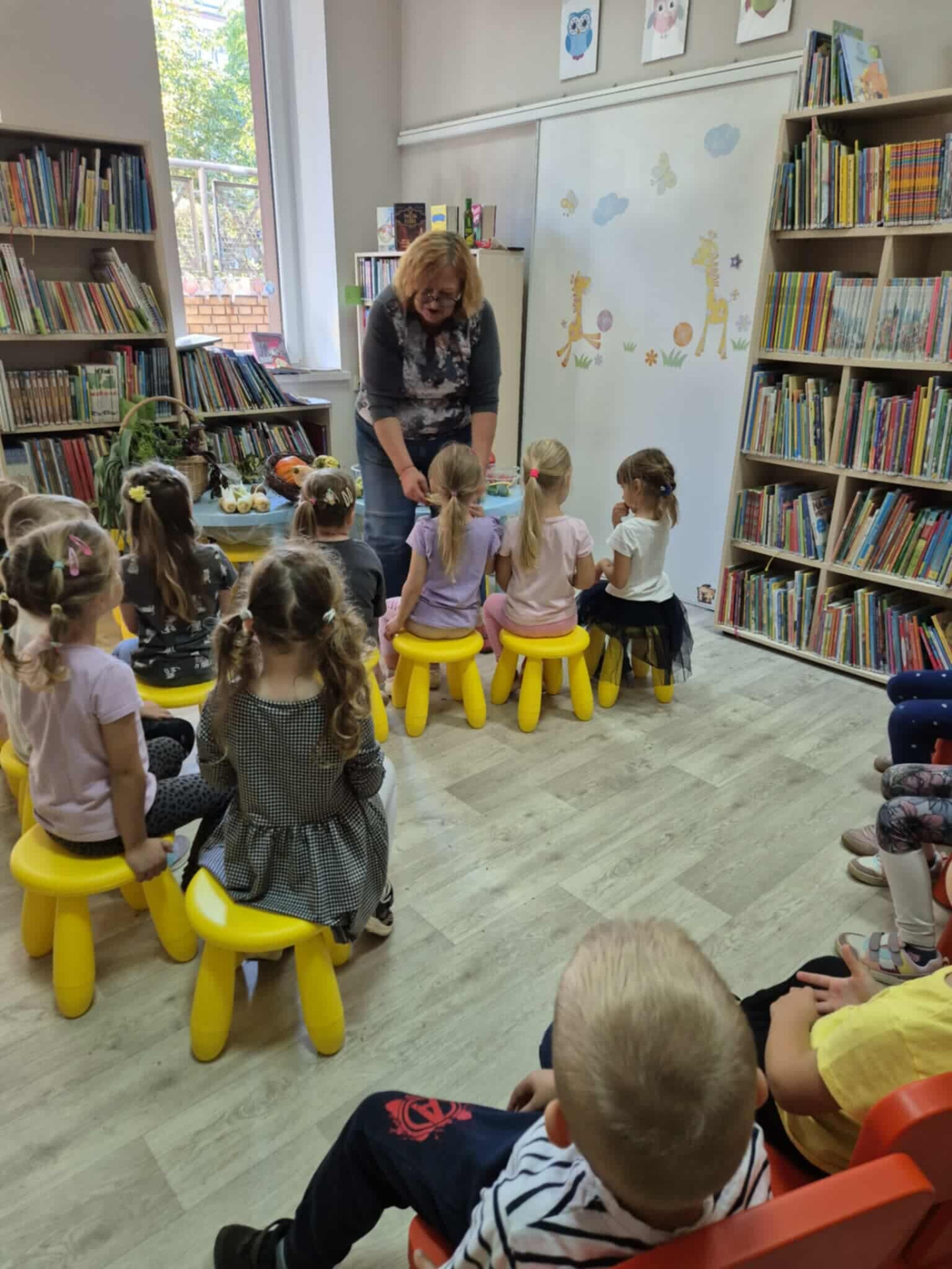 dzieci siedzące na żółtych tabroetach przed nimi dorosła kobieta