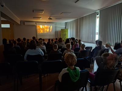 osoby dorosłe i dzieci siedzce w sali, gdzie na ścianie wyświela się film.