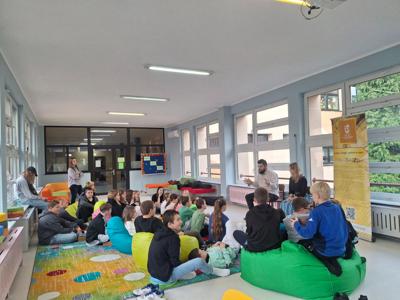 uczniowie siedzący na pufach i dywanie, przed nimi mężczyzna i kobieta trzymający instrumenty muzyczne