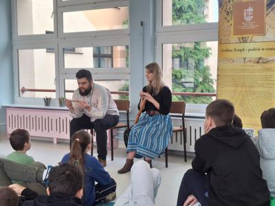 uczniowie siedzący na pufach i dywanie, przed nimi mężczyzna i kobieta trzymający instrumenty muzyczne