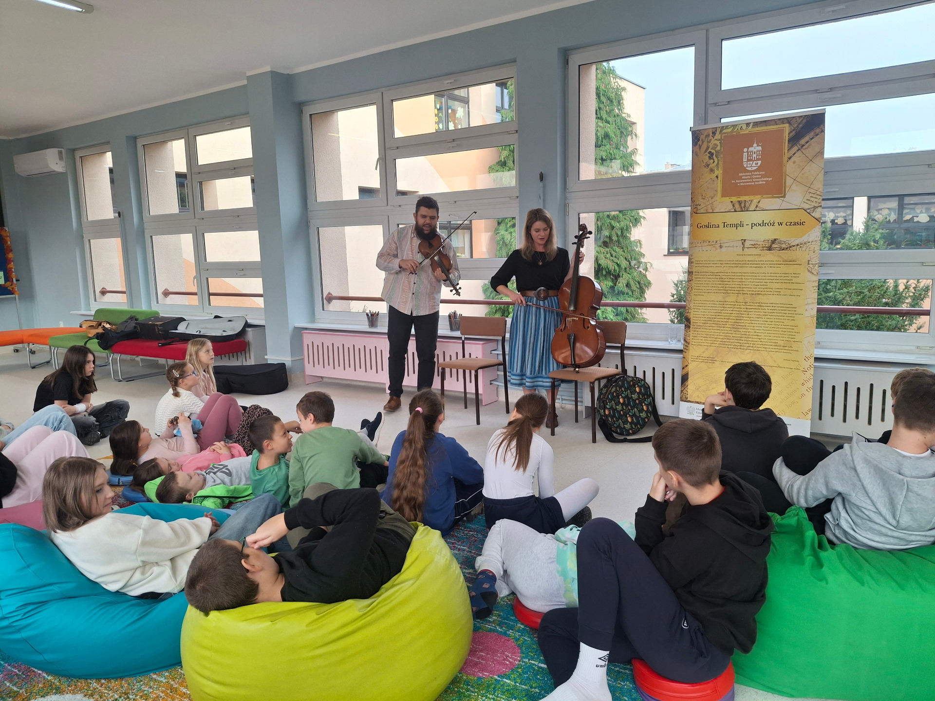 uczniowie siedzący na pufach i dywanie, przed nimi mężczyzna i kobieta trzymający instrumenty muzyczne