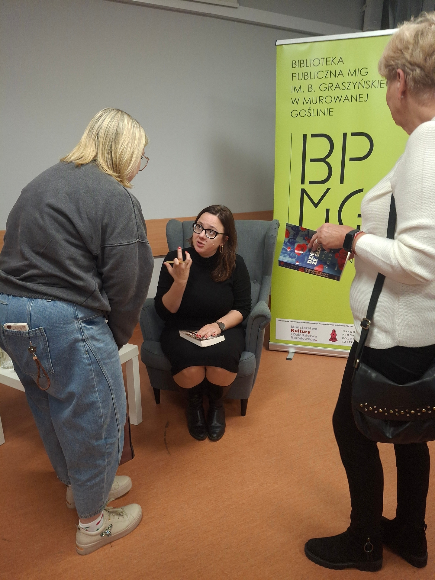 kobieta na fotelu podpsiuje książkę, przed nią dwie kobiety