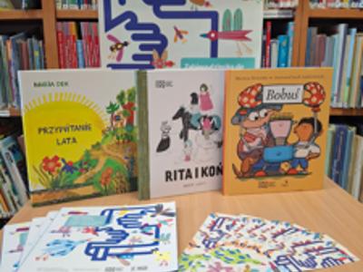 zdjęcie w pomieszczeniu bibliotecznym, tło plakat, na stole karty na naklejki, ksiązki, broszura kampani