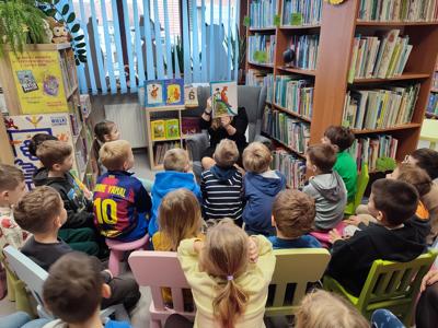 dzieci siedzące tyłem, osoba dorosła siedząca na fotelu z ksiązką w ręku