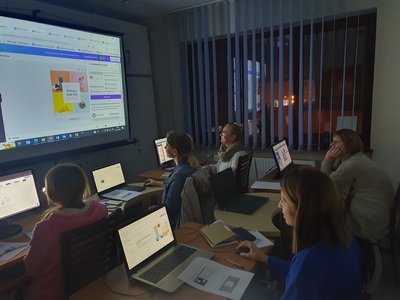 Kobiety siedzące przed monitorami komputerów. Przed biurkami duży ekran