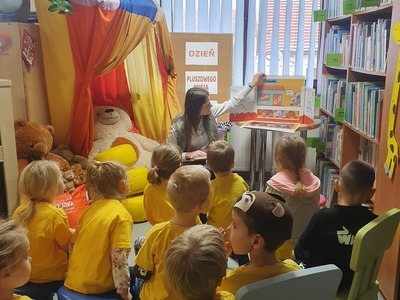 grupa dzieci w żółtych koszulkach siedzą na krzesłach i patrzą na kobietę, która pokazuje ilustracje