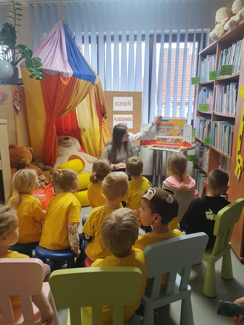 grupa dzieci w żółtych koszulkach siedzą na krzesłach i patrzą na kobietę, która pokazuje ilustracje