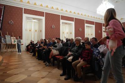 Publiczność oglądająca spektakl na krzesłach - przed sceną sztalugi z obrazami.