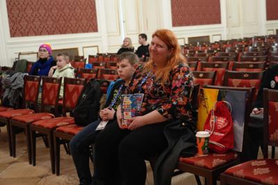 Garstka osób siedząca na publiczności. Na pierwszym planie uśmiechnięta pani trzymająca  puszkę WOŚP wraz z chłopczykiem.