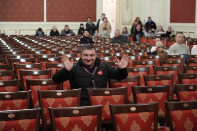 Garstka osób na widowni, pan z identyfikatorem wolontariusza WOŚP uśmiecha się i macha do kamery.