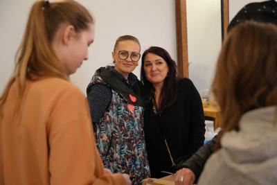 Dwie panie obejmują się i uśmiechają do kamery. Wokół nich ludzie.