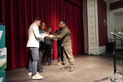 Grupa ludzi na scenie. Pan w mundurze wojskowym wręcza drugiemu panu podarunek. Za nimi czerwona kurtyna.