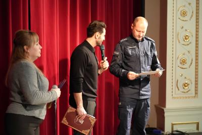 Trzy osoby na scenie : pani, pan z mikrofonem i pan w mundurze policyjnym. Za nimi czerwona kurtyna.