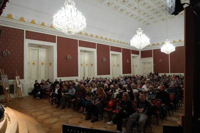Sala Kryształowa w Pałacu Książęcym w Żaganiu : schodki na scenę, publiczność siedząca na krzesłach, a nad nimi ogromne żyrandole.