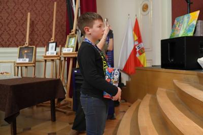 Chłopczyk z puszką na datki WOŚP stoi przed schodkami na scenę. Za nim obrazy na sztalugach oraz flaga Żagania.