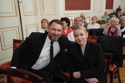 Ludzie siedzący na widowni. Na pierwszym planie elegancko ubrane, uśmiechnięte osoby obejmują się.