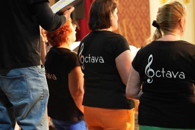 Grupa ludzi na scenie, występ chóru. Na koszulkach śpiewających znajduje się klucz wiolinowy i napis "octava".