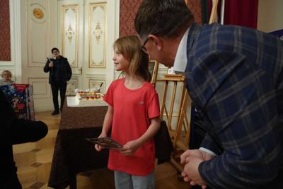 Pan nachyla się nad dziewczynką mówiąc coś do niej. W tle widać ogromne, zdobione drzwi Sali Kryształowej.