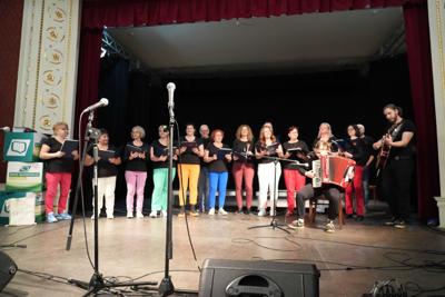 Grupa ludzi na scenie, występ chóru. Przed nimi dwie osoby, grające na akordeonie i gitarze.