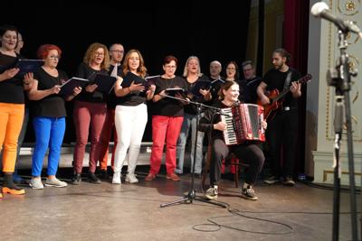 Grupa ludzi na scenie, występ chóru. Przed nimi dwie osoby, grające na akordeonie i gitarze.