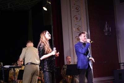 Dwoje artystów śpiewa na scenie, za nimi orkiestra wojskowa.