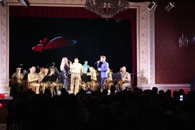 Dwoje artystów śpiewa na scenie, za nimi orkiestra wojskowa.