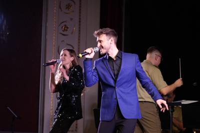 Dwoje artystów śpiewa na scenie, za nimi dyrygent orkiestry wojskowej.