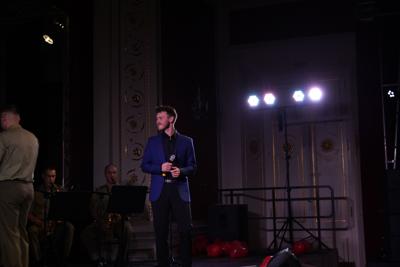 Pan z mikrofonem stoi na scenie. Za nim orkiestra wojskowa.