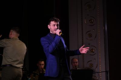 Pan śpiewa na scenie. Za nim orkiestra wojskowa.