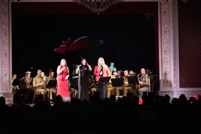 Trzy artystki śpiewają na scenie. Za nimi orkiestra wojskowa.