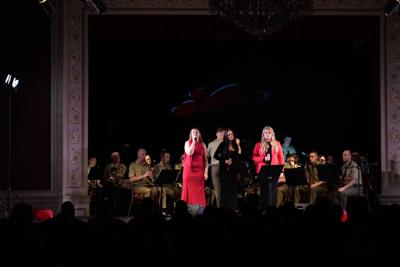 Trzy artystki śpiewają na scenie. Za nimi orkiestra wojskowa.