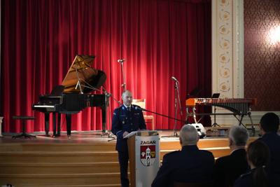 Pan przy mównicy przemawia do publiczności, za nim scena z instrumentami muzycznymi.