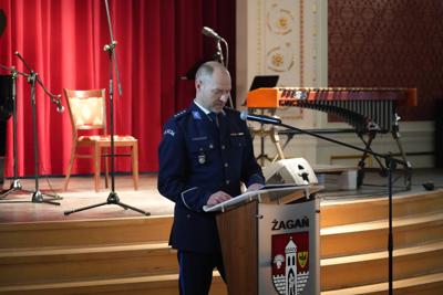 Pan przy mównicy przemawia, za nim scena z instrumentami muzycznymi.