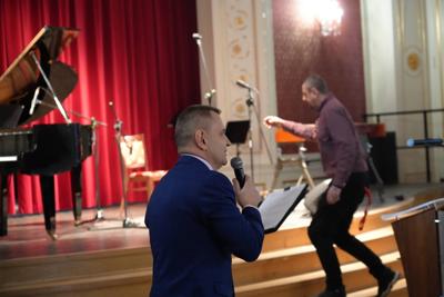 Pan z mikrofonem przemawia, za nim pan wchodzi na scenę z instrumentami muzycznymi.