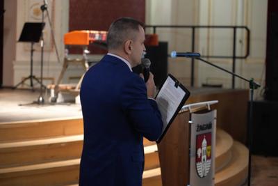 Pan z mikrofonem przemawia, za nim scena z instrumentami muzycznymi.