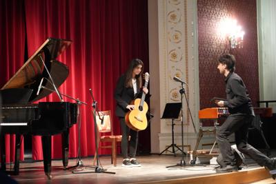 Dziewczyna stoi na scenie z gitarą, na scenę wchodzi pan. Wokół stoją inne instrumenty.