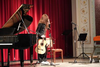 Dziewczyna stoi z gitarą na scenie. Wokół stoją inne instrumenty.
