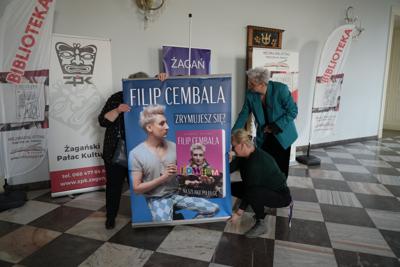 Grupa osób ustawia plakat z okazji spotkania autorskiego z Filipem Cembalą.