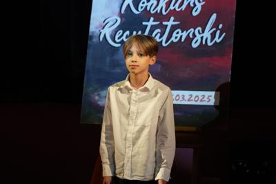 Chłopiec recytuje dla publiczności, za nim plakat konkursu recytatorskiego.